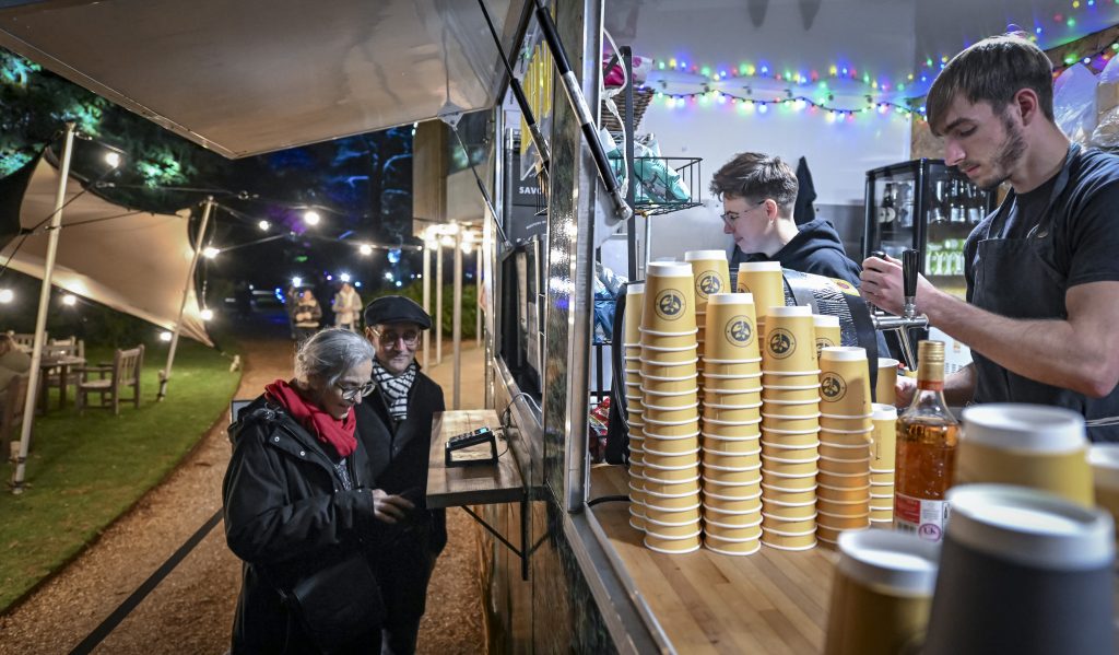 In the centre of the image, two visitors are making a purchase from two baristas who are standing in a trailer that is lit with rainbow fairy lights. On the counter are several 