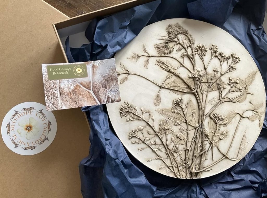 A Hope Cottage Botanical Arts product, featuring a beautifully crafted clay plate with intricate botanical engravings, alongside an open box with a label visibly displayed.