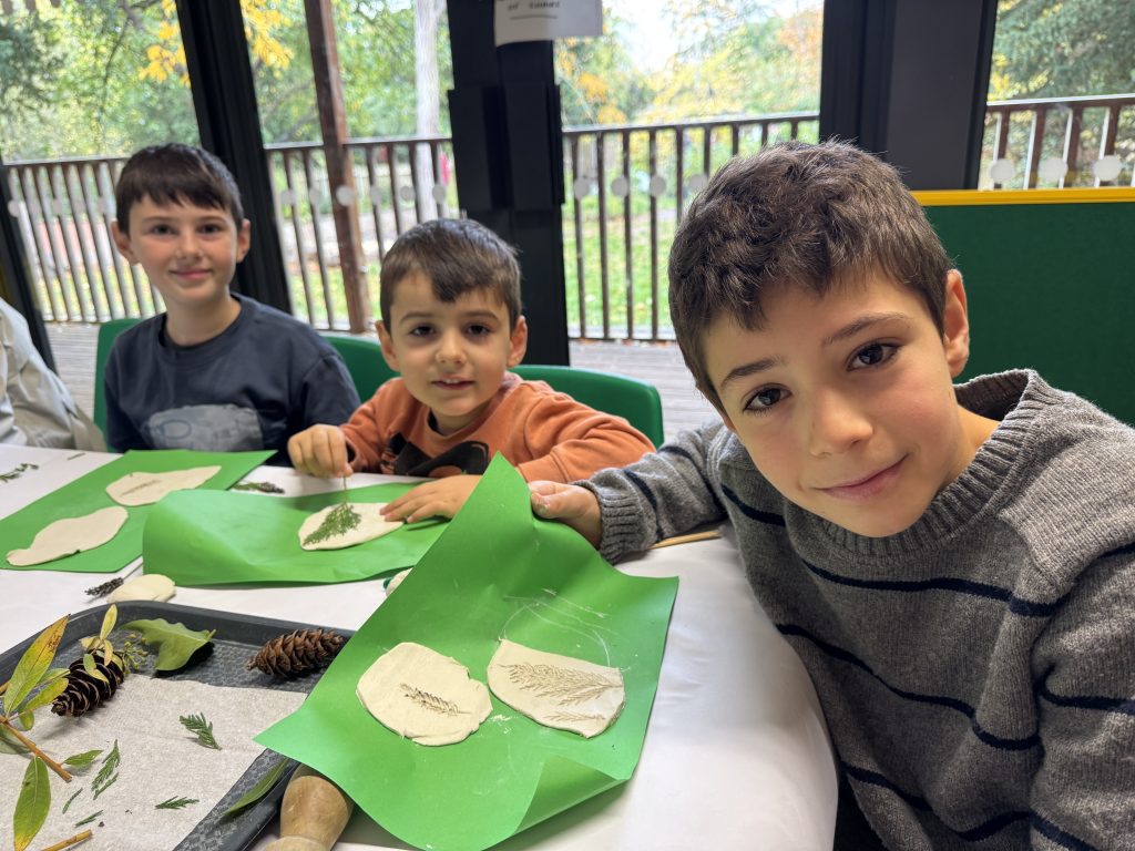 Three children engaged in a craft activity, sitting at a table with papers and natural items like leaves and pine cones. They are smiling and looking at the camera in a brightly lit room with windows showing a view of trees.