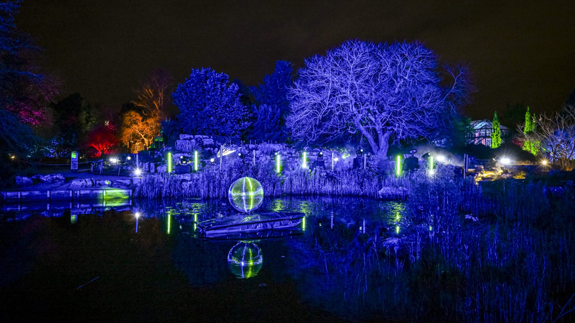A disco ball sits on a small boat in the middle of a lake surrounded by illuminated trees and shrubs.
