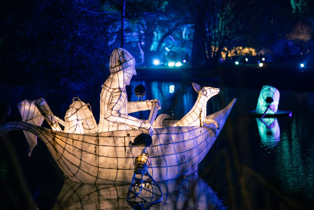 Illuminated sculpture of a person rowing a boat with a dog figure at the front, floating on a pond at night, surrounded by blue and teal lights reflected in the water.
