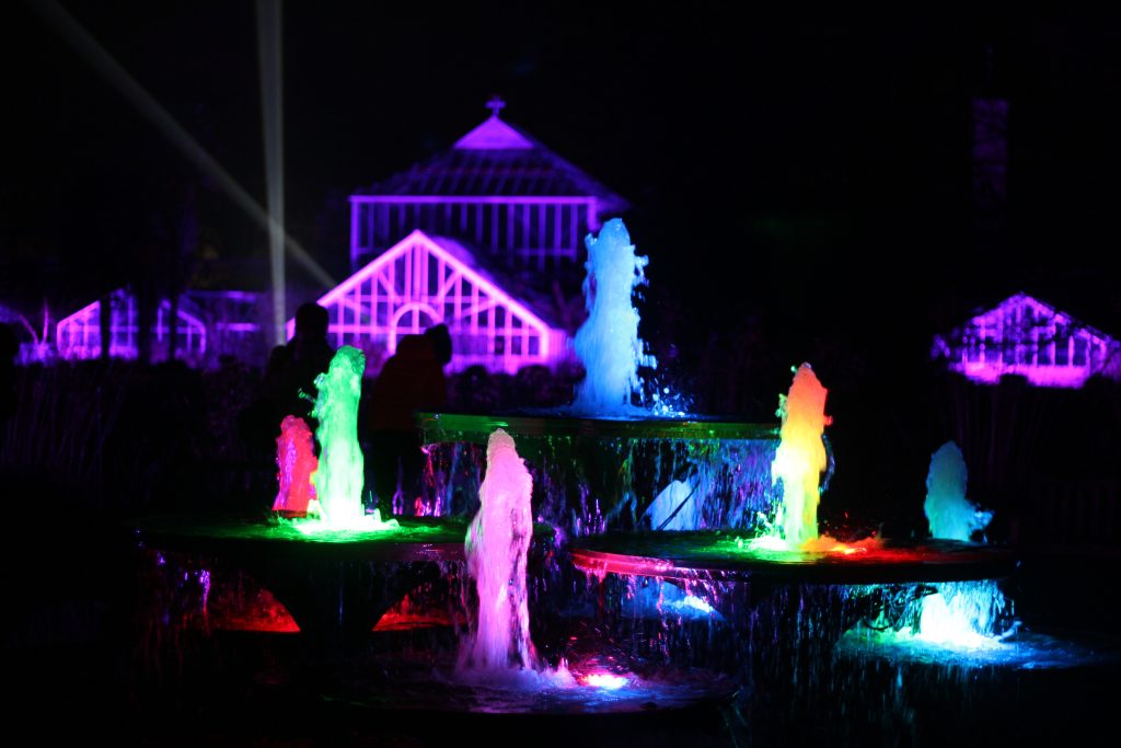 A water fountain at night, lit up with multicoloured lights in front of an greenhouse illuminated in purple.