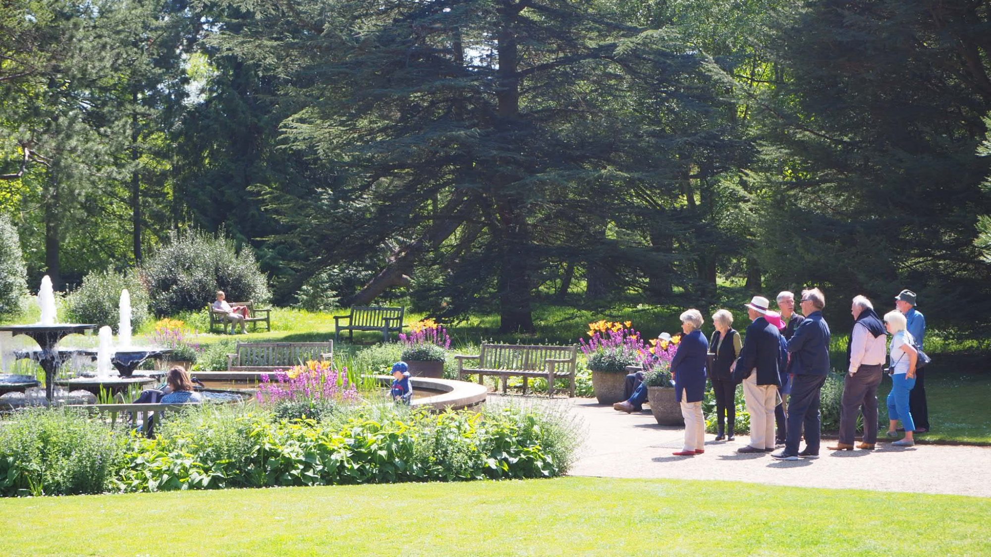 A group of people standing on a pathway observes a large fountain in a lush, sunlit garden. The scene includes vibrant flowers, well-maintained lawns, and tall trees. Benches are scattered around the garden.