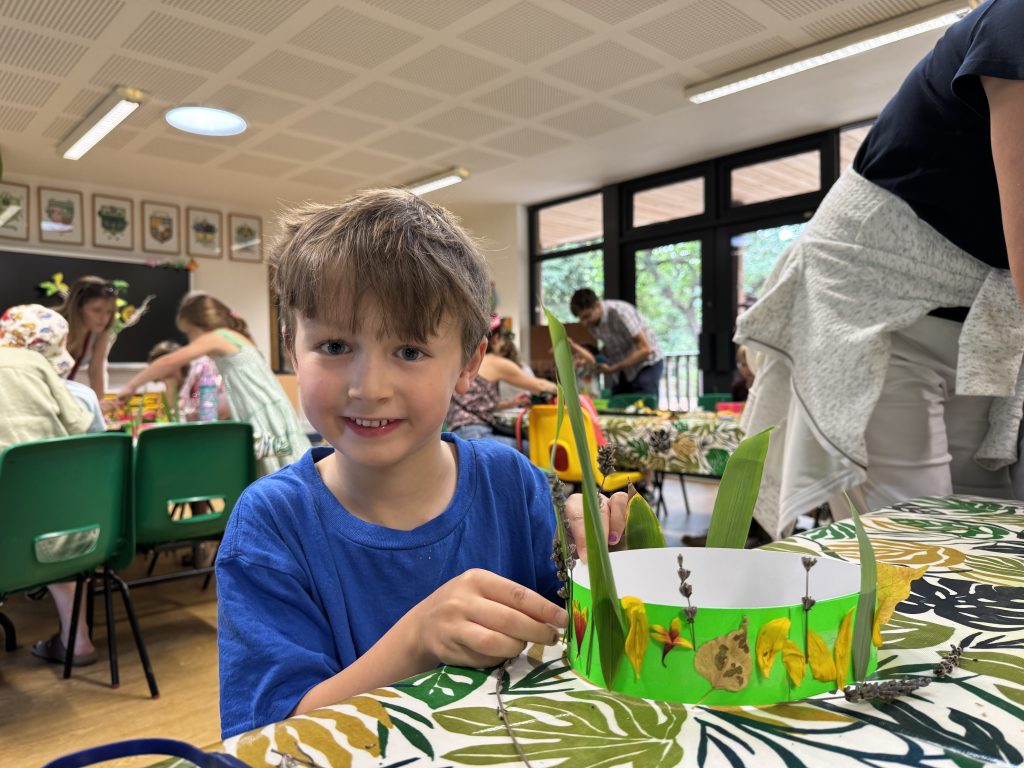 A child smiling while crafting at a table with a leafy crown, surrounded by others in a classroom setting. Vibrant decorations and artwork are visible.