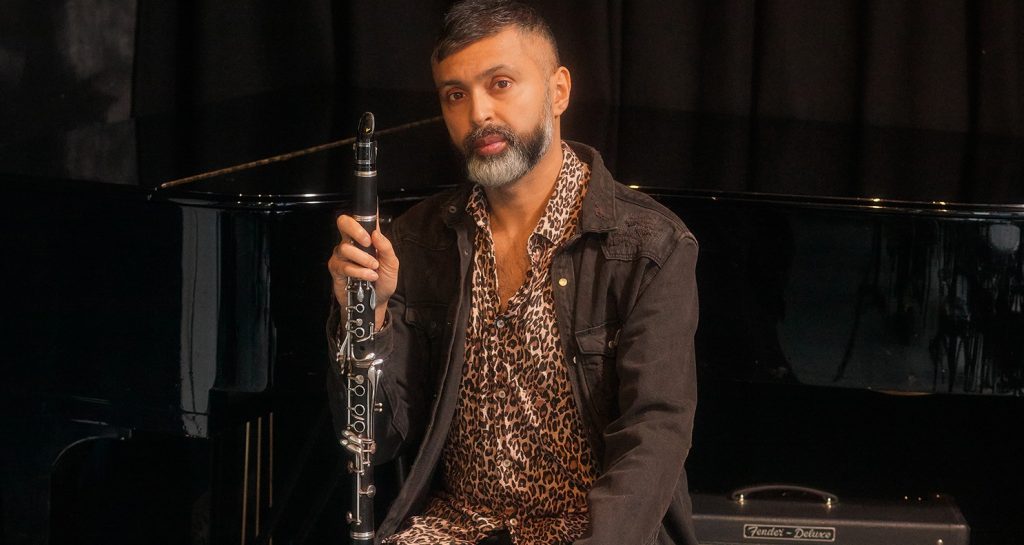 A person is seated with a clarinet, in front of a piano. They are wearing a leopard print outfit and have a short beard.