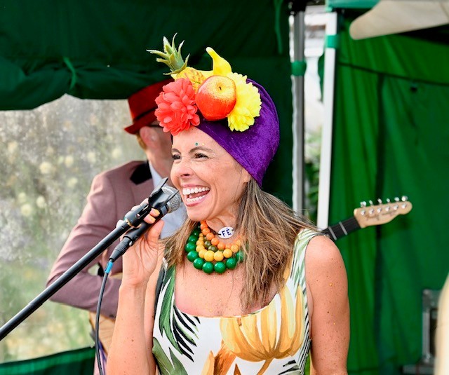 A woman singing into a microphone wears bright bead necklaces, a head scarf with flowers and plastic fruit attached. A guitarists is slightly obscured in the background.
