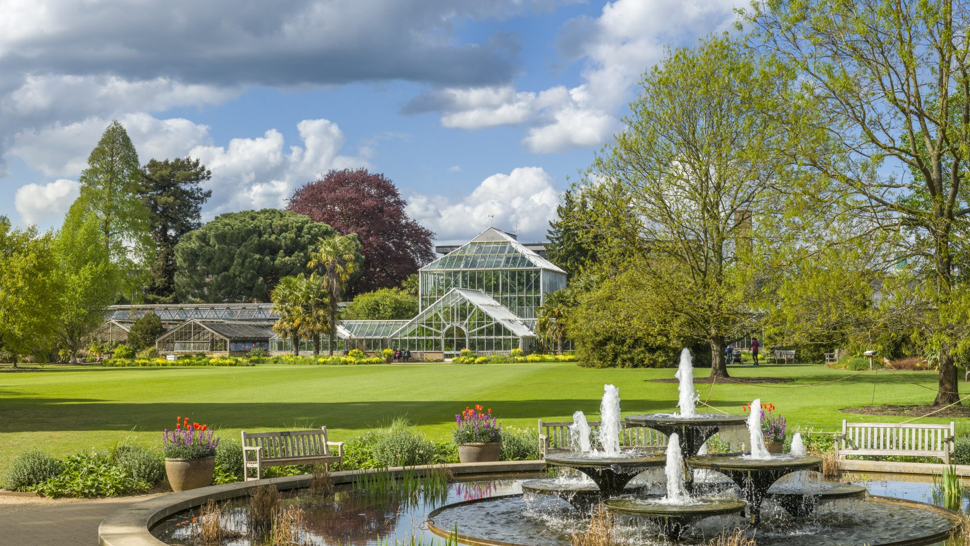 The image shows the glasshouse and surrounding gardens at Cambridge University Botanic Garden, featuring a clear blue sky with some clouds. In the foreground, there is a circular fountain surrounded by benches and decorative flower pots. The landscape is lush with various trees and well‑maintained lawns.