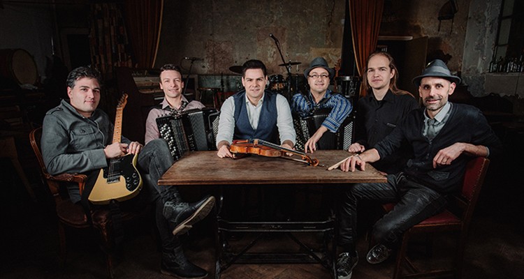 A group of six individuals is seated around a wooden table in a dimly lit room, with musical instruments like a guitar, violin, and accordion visible. There is a drum set in the background.