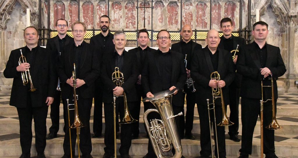 A group of eleven people dressed in black stands in a grand, ornate interior holding brass instruments. The setting features intricate stonework and arched designs.