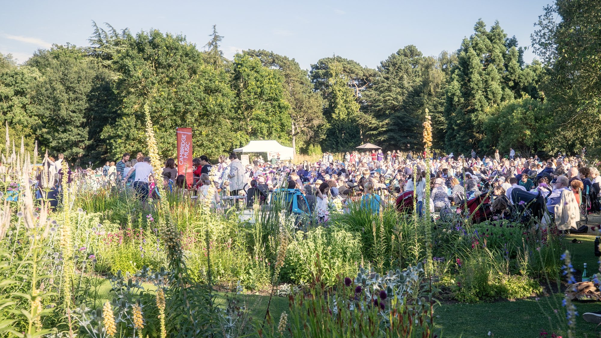 A large outdoor gathering in a lush, green space with numerous people seated on chairs or blankets. The area is surrounded by vibrant plants and trees under a clear blue sky. In the background, there are small tents and banners, indicating an organized event.