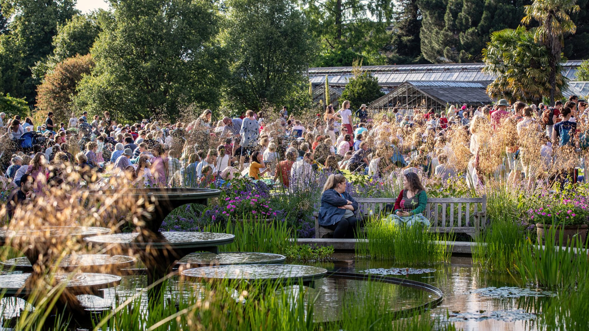 A large crowd gathers in a lush garden setting, filled with various plants and flowers. In the foreground, a water feature is visible. Some people are seated on benches, engaging in conversation. The scene is lively and bathed in natural light.