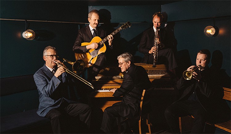 Five individuals are playing musical instruments in a dimly lit room. They are seated with a guitar, saxophone, trombone, trumpet, and piano. The setting appears to be a jazz band performance area.