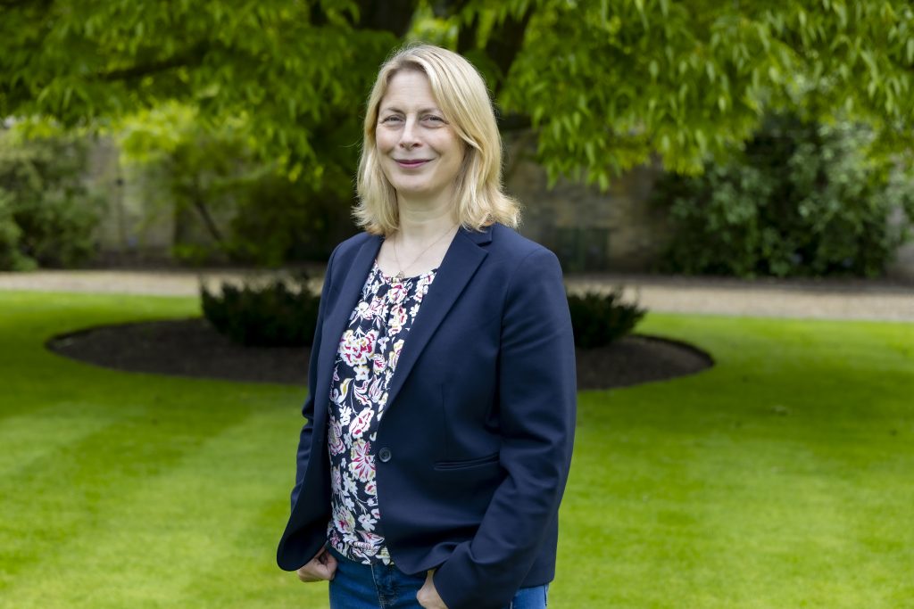 A woman is standing and smiling in a park with green grass and a tree and bushes blurred in the background.