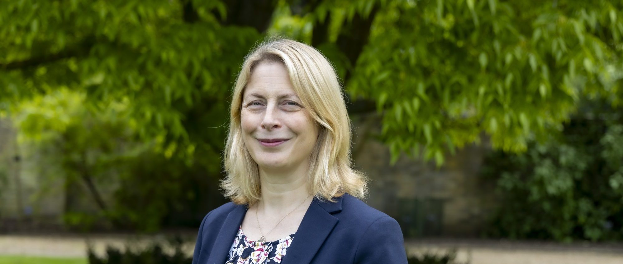A woman is standing and smiling in a park with green grass and a tree and bushes blurred in the background.