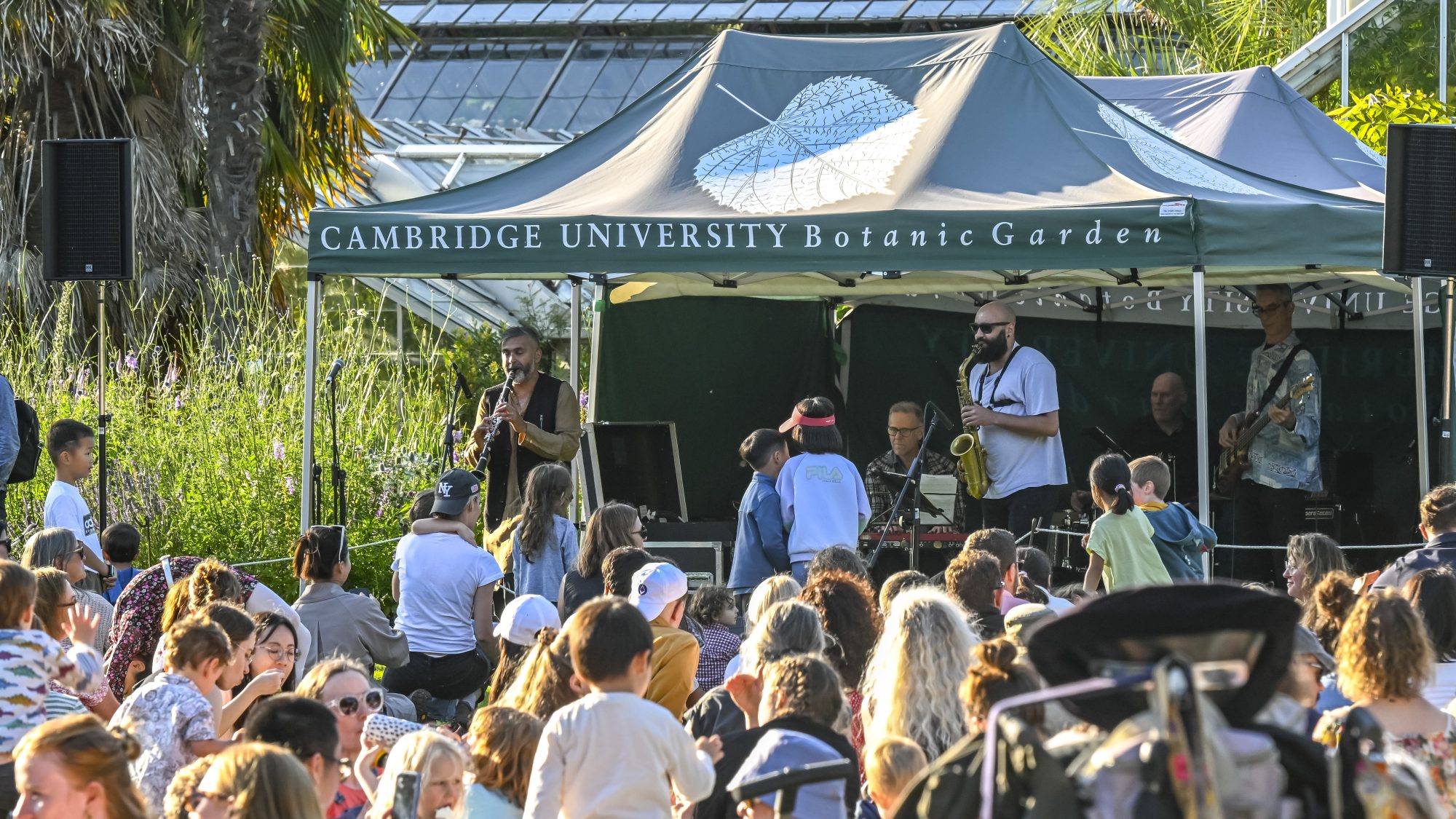Outdoor event at Cambridge University Botanic Garden with a musical performance under a tent. People are gathered on the grass, enjoying the event amidst greenery and a sunny atmosphere.