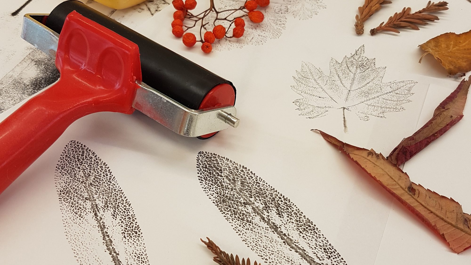 A table displays art supplies and nature-themed prints. There's a red roller, sheets with leaf prints, various dried leaves and twigs, and several colourful ink pads.
