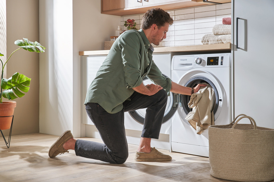 A person crouches in front of a washing machine, transferring laundry from the machine to a rope laundry basket.