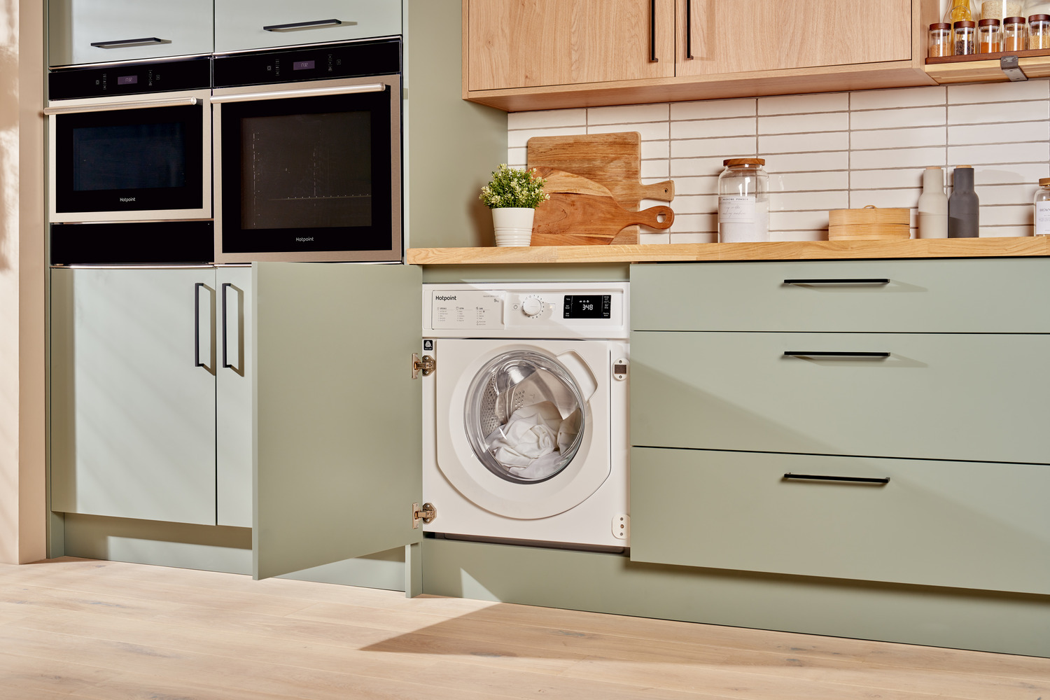 A white Hotpoint washing machine is integrated into a green kitchen cabinet. The washing machine door is open, showing clothes inside.