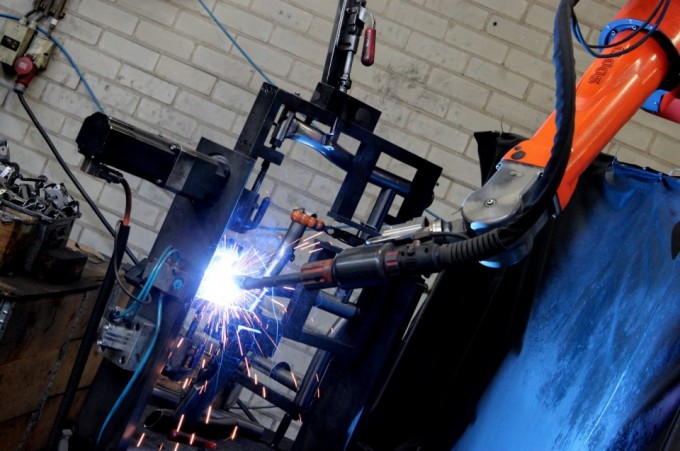 Auf der einen Seite befestigt ein Mitarbeiter einen Rahmen in einer dafür vorgesehenen Halterung und auf der anderen Seite schweißt der Roboter. Im Anschluss werden die Seiten gewechselt. Der Mitarbeiter prüft den fertigen Rahmen und der Roboter schweißt den Nächsten.