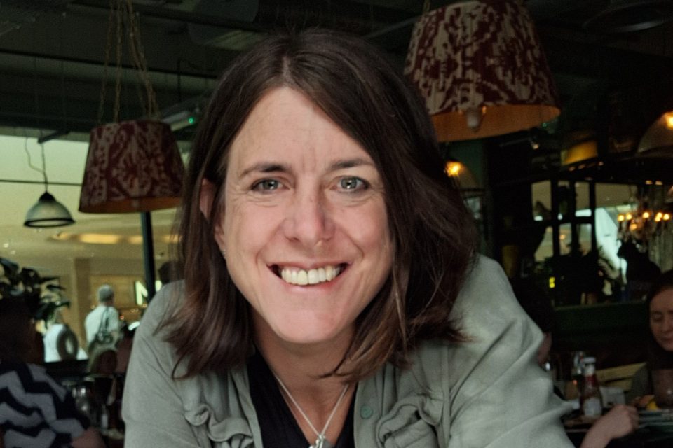 Helen is smiling, leaning towards the camera while sitting in a restaurant.