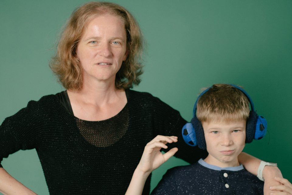 A mother and son face the camera with serious expressions on their face. The son is wearing ear defenders.