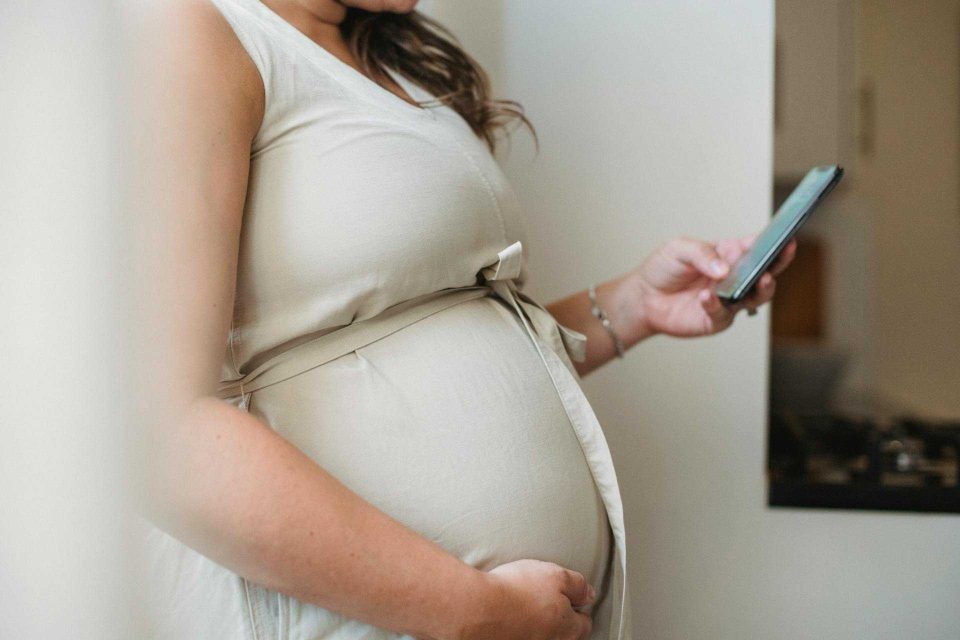 A pregnant woman looks at her phone, with one arm resting on her stomach.
