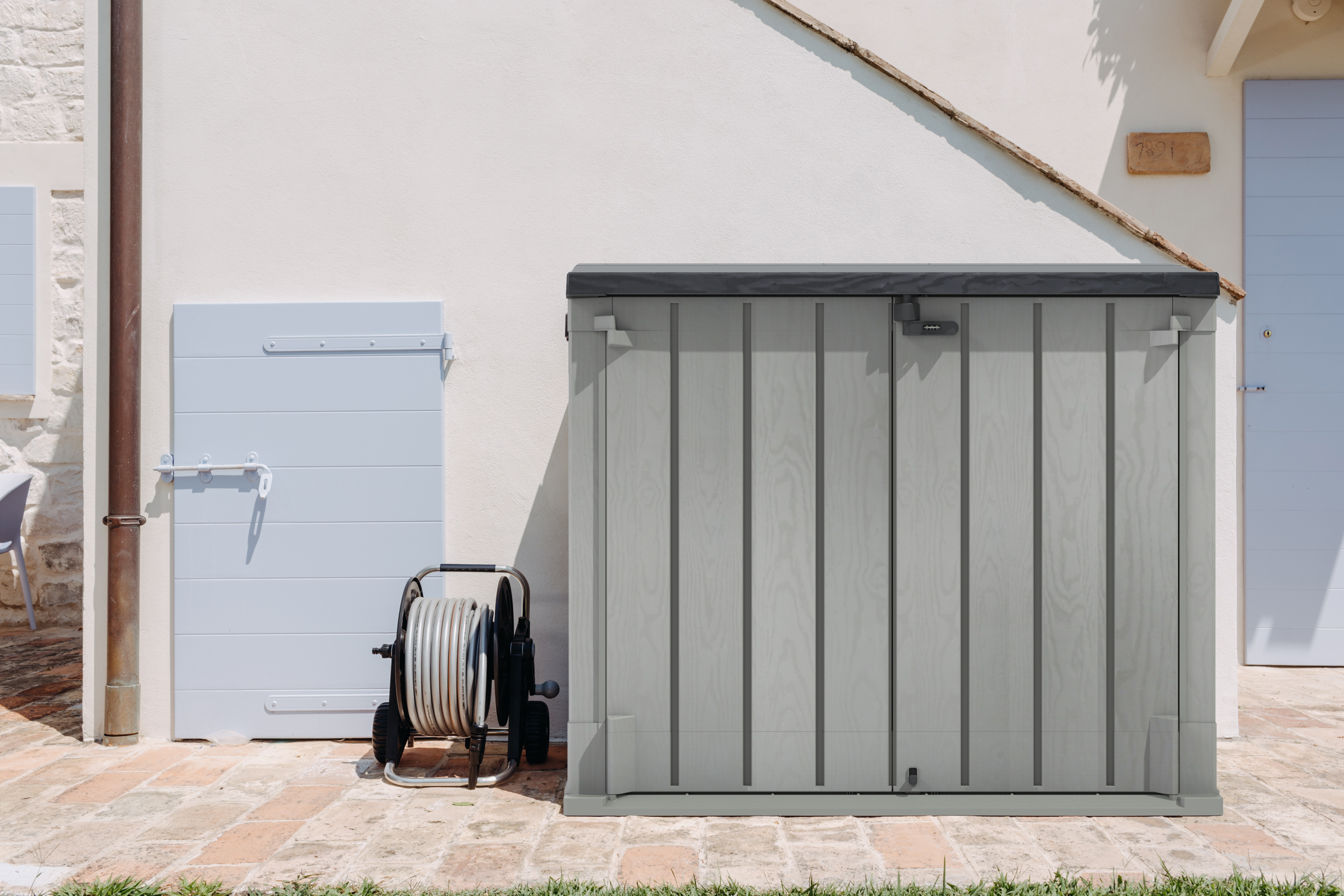 A plastic garden storage box situated by the outside side of a house with a ladder above it.
