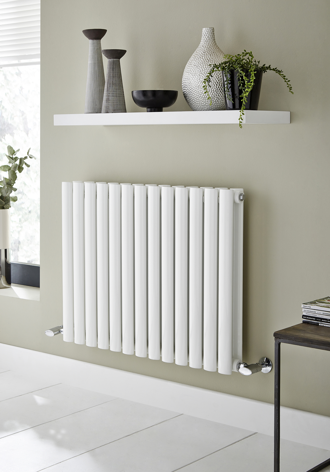 White radiator mounted on a wall in a modern interior setting with vases and decorative elements hung above it.