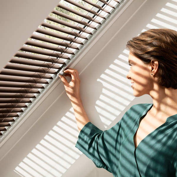 A VELUX window with a woman opening the blinds on a bright sky outside. 