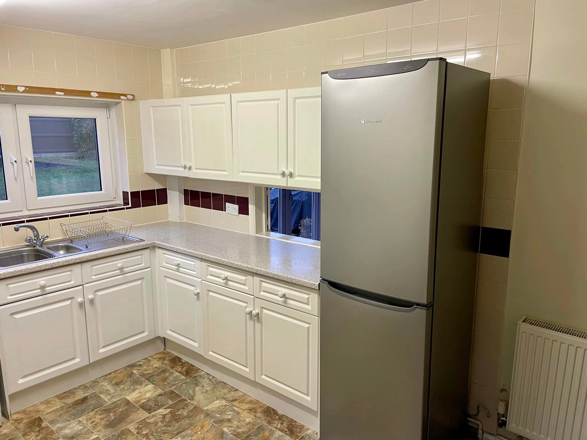 A tired, dull looking kitchen with white cupboards and metallic fridge.