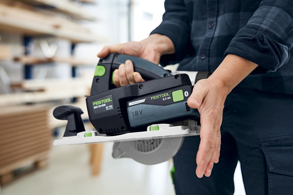Eine Person hält die Festool HKC 55 K Akku-Handkreissäge mit einer Hand am Griff und der anderen stabilisierend an der Grundplatte. Im Hintergrund sieht man eine helle, aufgeräumte Werkstatt.