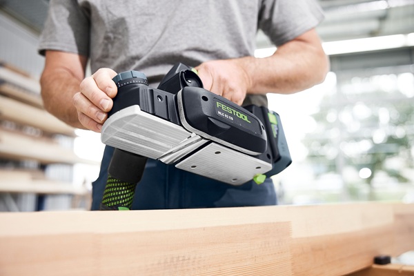 Ein Handwerker bearbeitet ein Holzstück mit dem Akku-Hobel Festool HLC 82 EB. Die Arbeitsumgebung ist eine helle, moderne Werkstatt.