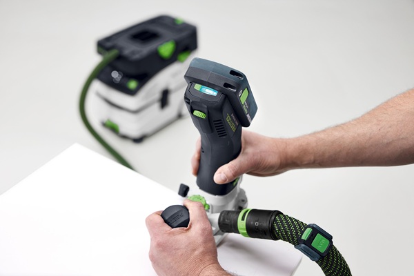 Nahaufnahme der Akku-Kantenfräse Festool MFKC 700 KA im Einsatz. Ein Anwender führt die Maschine mit beiden Händen entlang einer weißen Werkstückkante. Die Fräse ist mit einem Absaugschlauch verbunden, im Hintergrund befindet sich der zugehörige mobile Staubsauger.