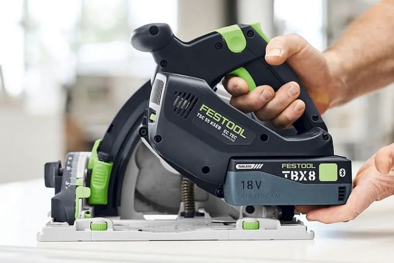 Akkubetriebene Festool Tauchsäge TSC 55 mit 18-Volt-Akkupack, auf einem Tisch aufgesetzt, wird von einer Person mit beiden Händen bedient; Detailansicht in Werkstattumgebung.