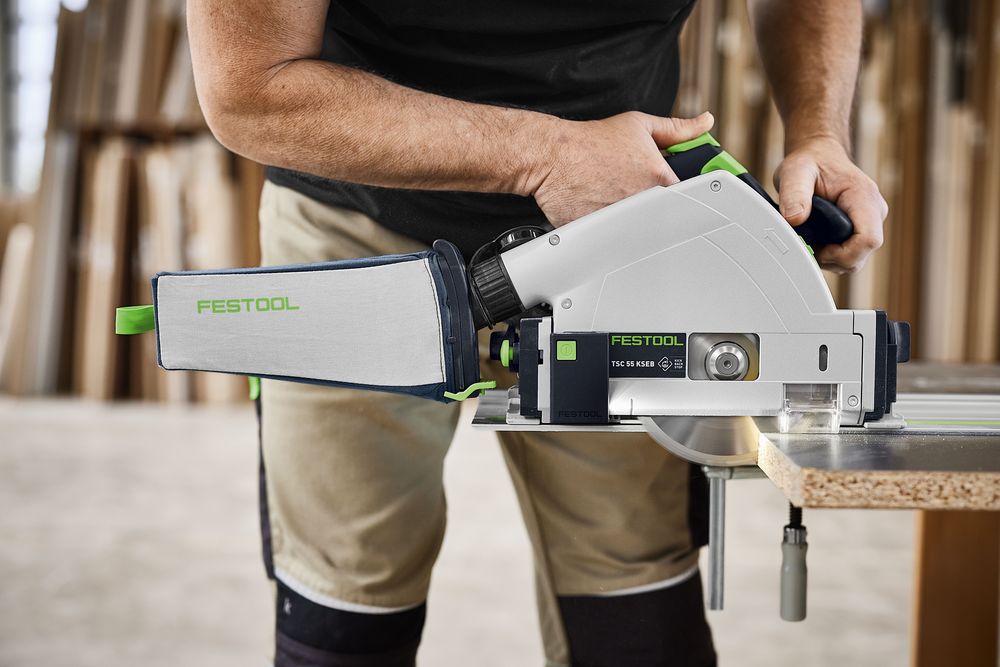 Festool Akku-Tauchsäge TSC 55 KSEB mit Staubfangbeutel schneidet eine Holzplatte entlang der Kante; Detailansicht der Maschine im Einsatz in einer Werkstatt, gehalten von einer Person.