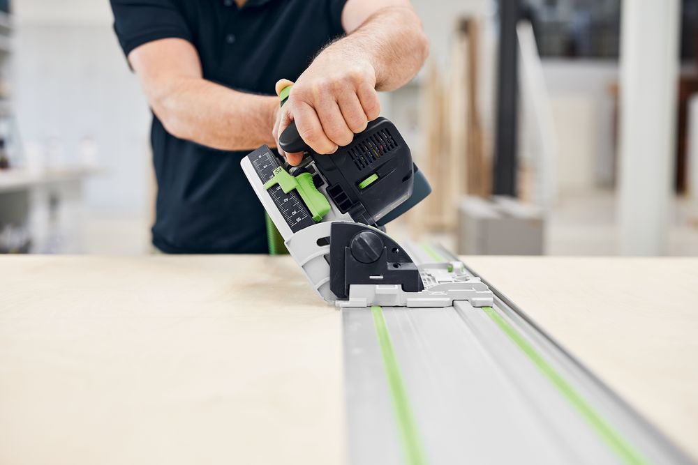 Person führt eine Festool Akku-Tauchsäge TSC 55 KSEB auf einer Führungsschiene und schneidet präzise eine Holzplatte; Ansicht von vorne mit Fokus auf Säge und Schiene in Werkstattumgebung.