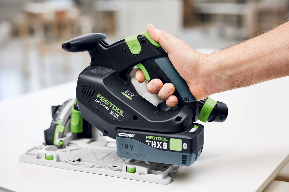 Hand hält eine Festool Akku-Tauchsäge TSC 55 KSEB mit 18-Volt-Akkupack auf einer Holzplatte; Detailansicht der Maschine von der Seite in heller Werkstattumgebung.