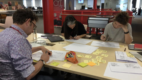 Three people gathered around a table with their heads down, sketching.