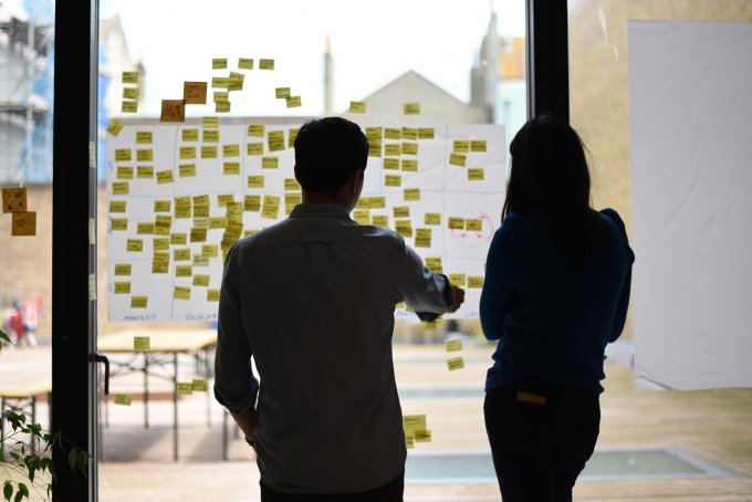 Two people having a discussion in front of a wall of post-it notes.