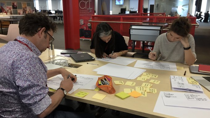 Three people gathered around a table with their heads down, sketching.
