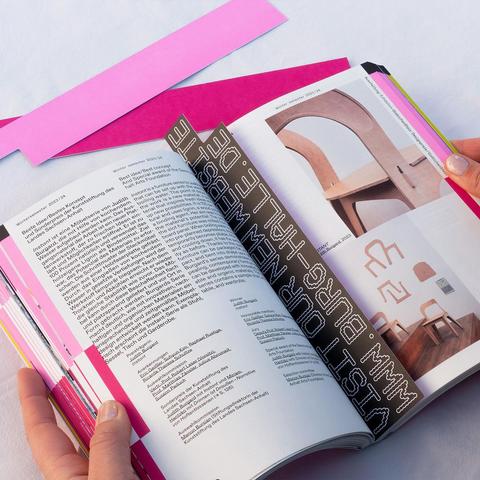 Open book held by hands showing text in German and photos of wooden chairs with curved backs