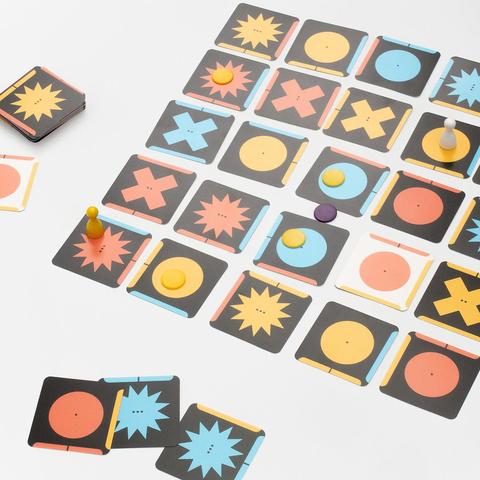 Board game with square cards showing colored shapes and symbols arranged in a grid with small round and pawn-shaped game pieces on some cards