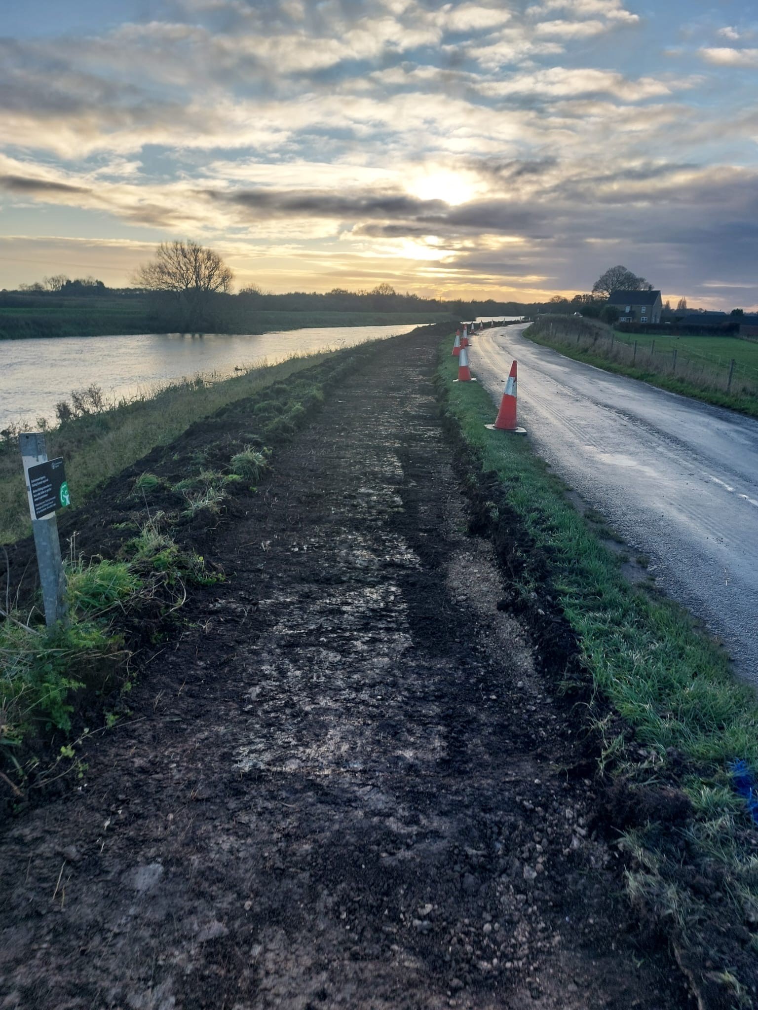 Traffic cones and recent ground works along a river embankment at sunset.