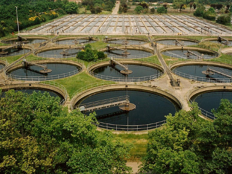 Aerial view of a wastewater treatment facility with multiple circular tanks surrounded by greenery and infrastructure.