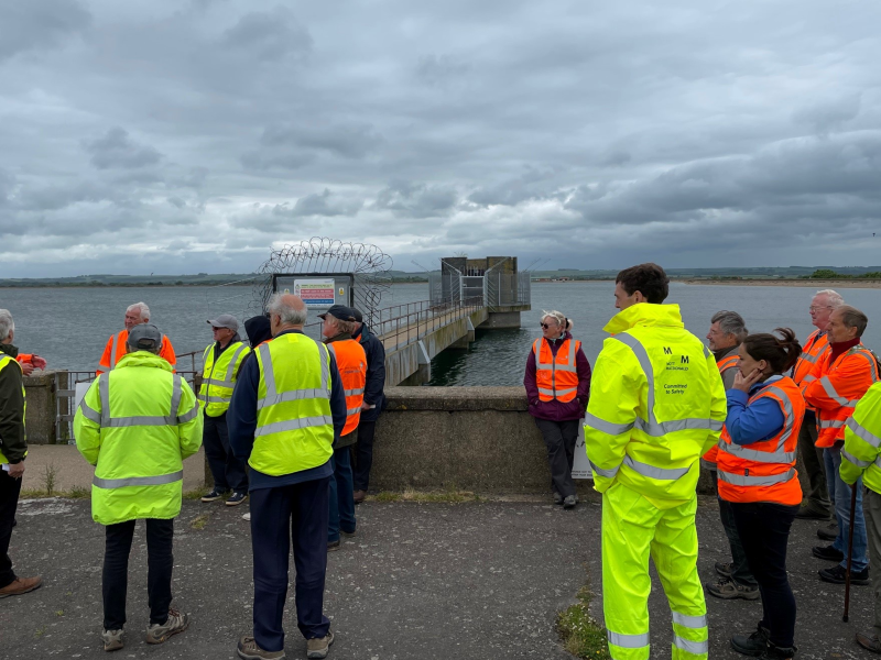 A group of people in high-visibility jackets gathers by a waterway, with a jetty and cloudy sky in the background.