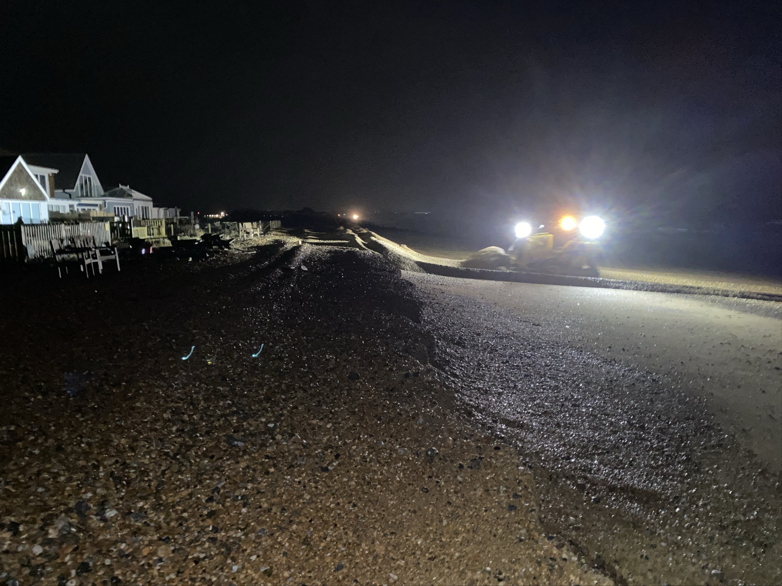 Nighttime view of Pevensey Beach showing beach erosion and wet shingle. Bright lights from a bulldozer illuminate the work area, with houses visible along the left side. The scene highlights ongoing beach restoration efforts after severe weather.