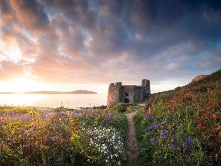 A narrow, wild flower lined footpath leads to the Castel Fort on Pendennis Head, while a dramatic dawn breaks over the sea beyond.