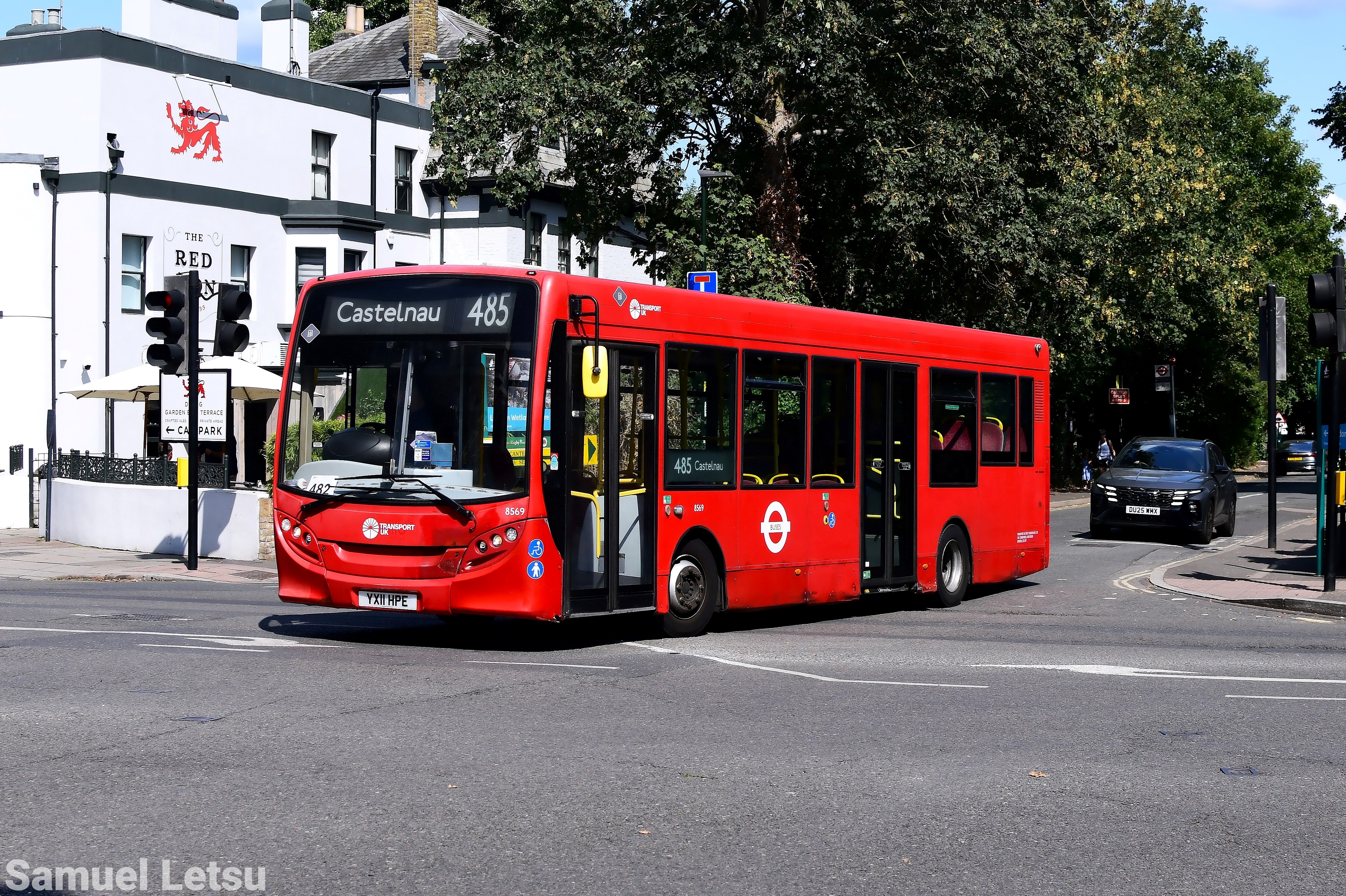 Single-decker red bus on route 485 to Castelnau turning at an intersection, passing a white building with traffic lights and another vehicle in the background