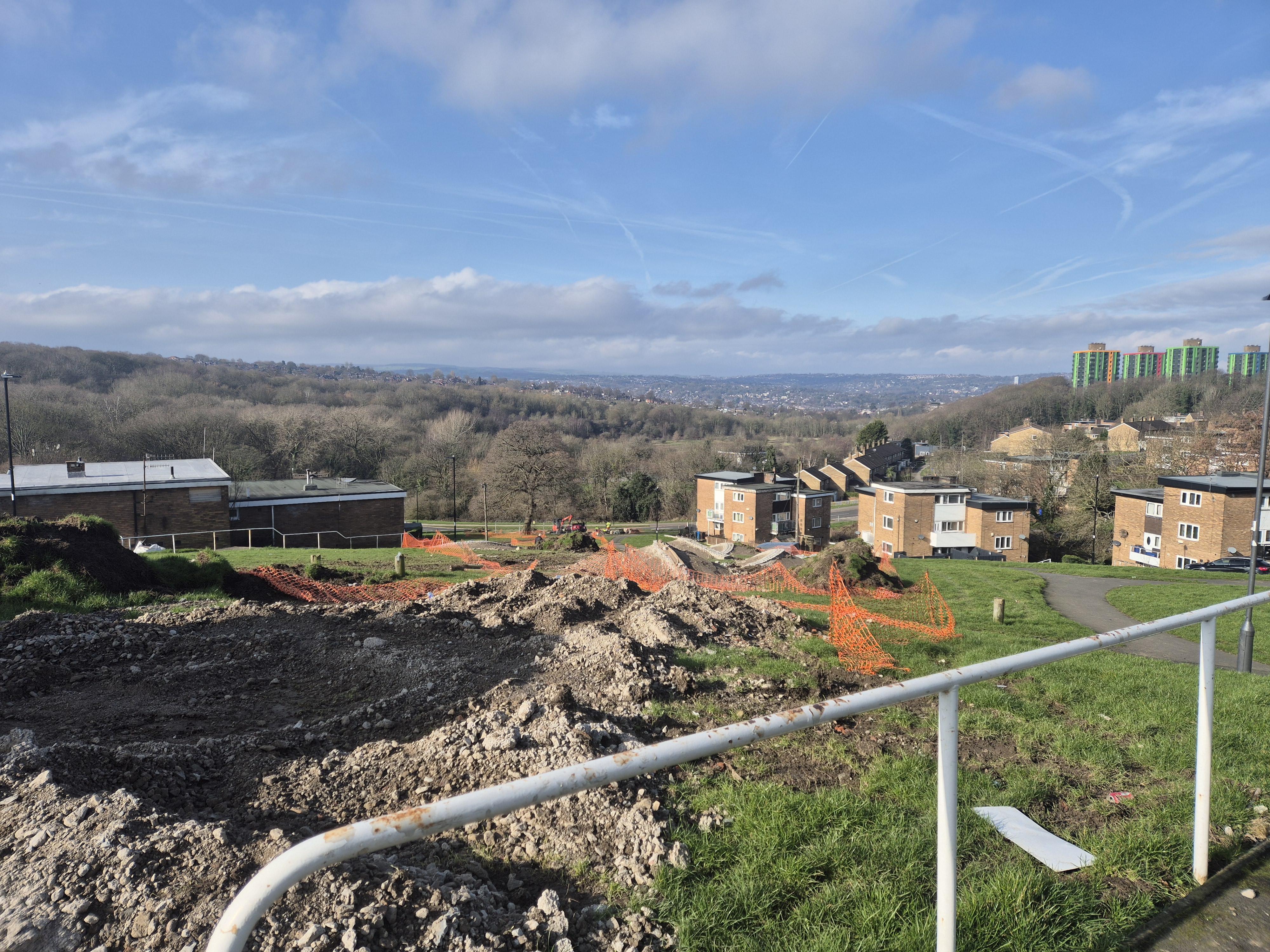 View from the top of Spotswood Mount, looking down over mounds of overturned earth. Orange plastic safety net fencing surrounds the mounds and there are houses and trees in the distance.