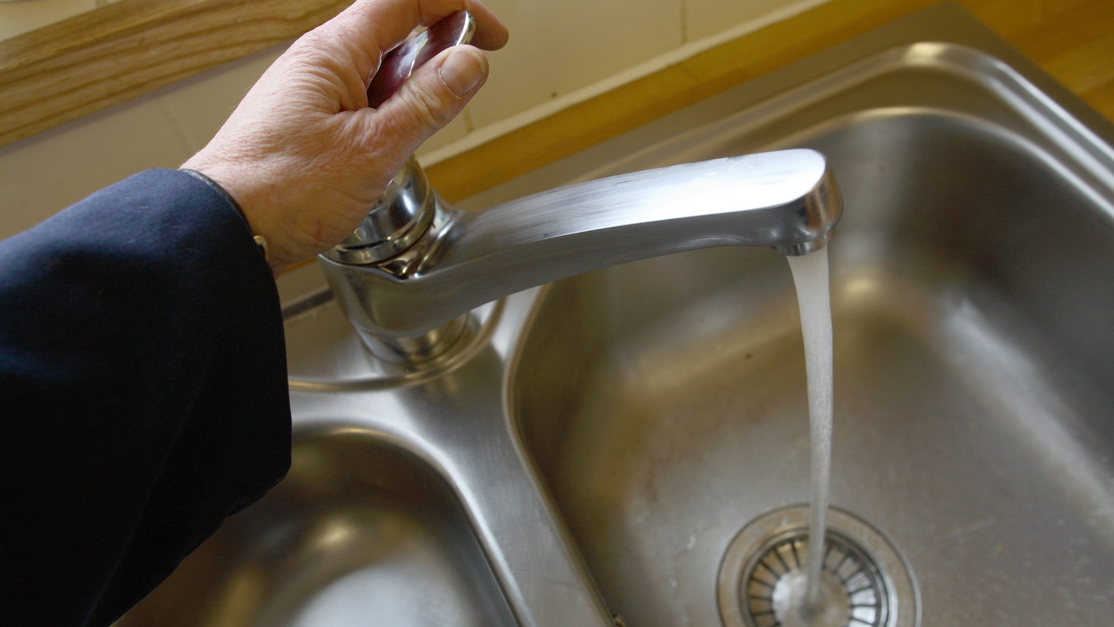 A hand operates a modern stainless steel kitchen faucet, with clear water flowing into a shiny metal sink.